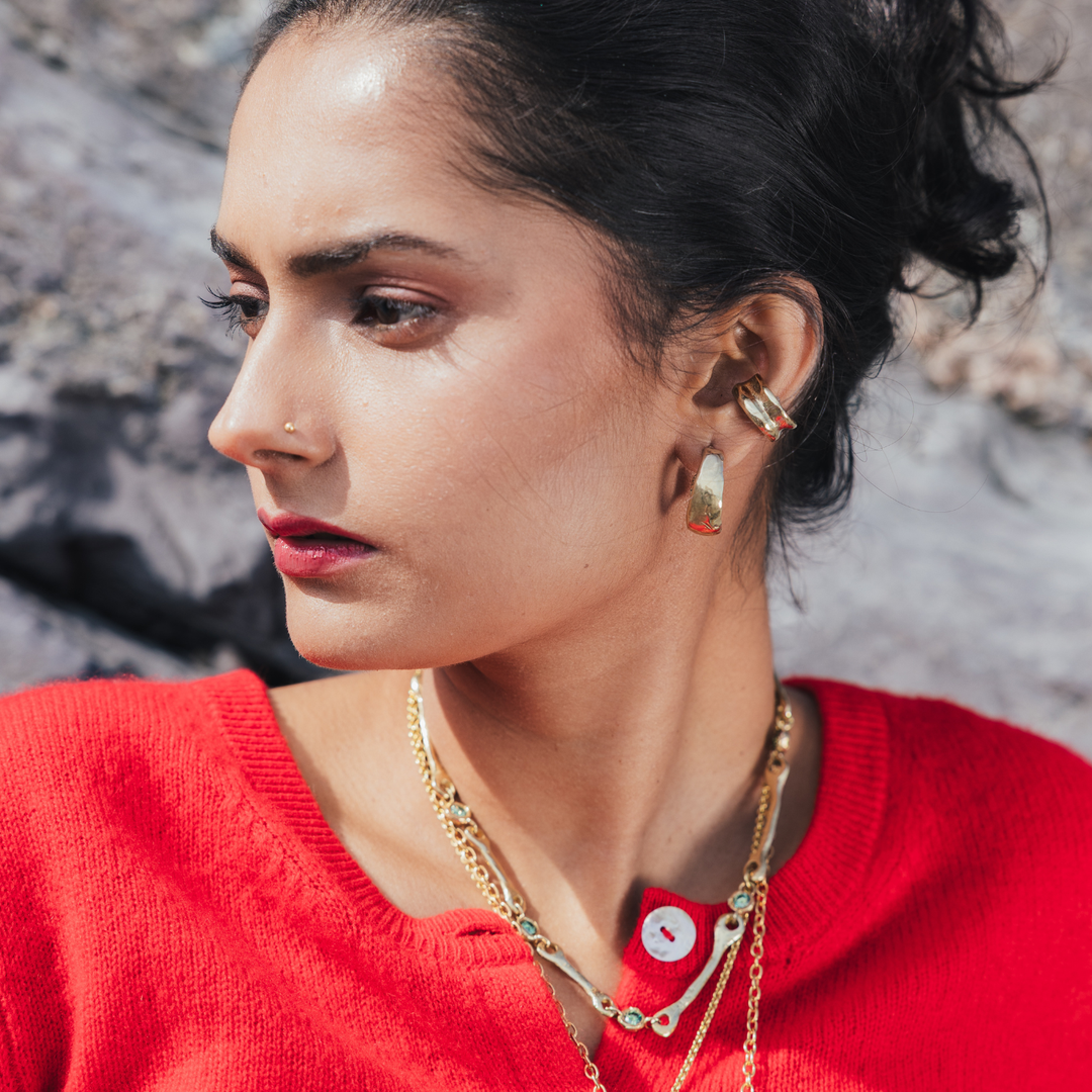 A close-up lifestyle shot of a woman with dark hair in a bun, wearing gold layered necklaces and earrings, a red knit top, and a nose ring, set against a blurred gray rocky background.