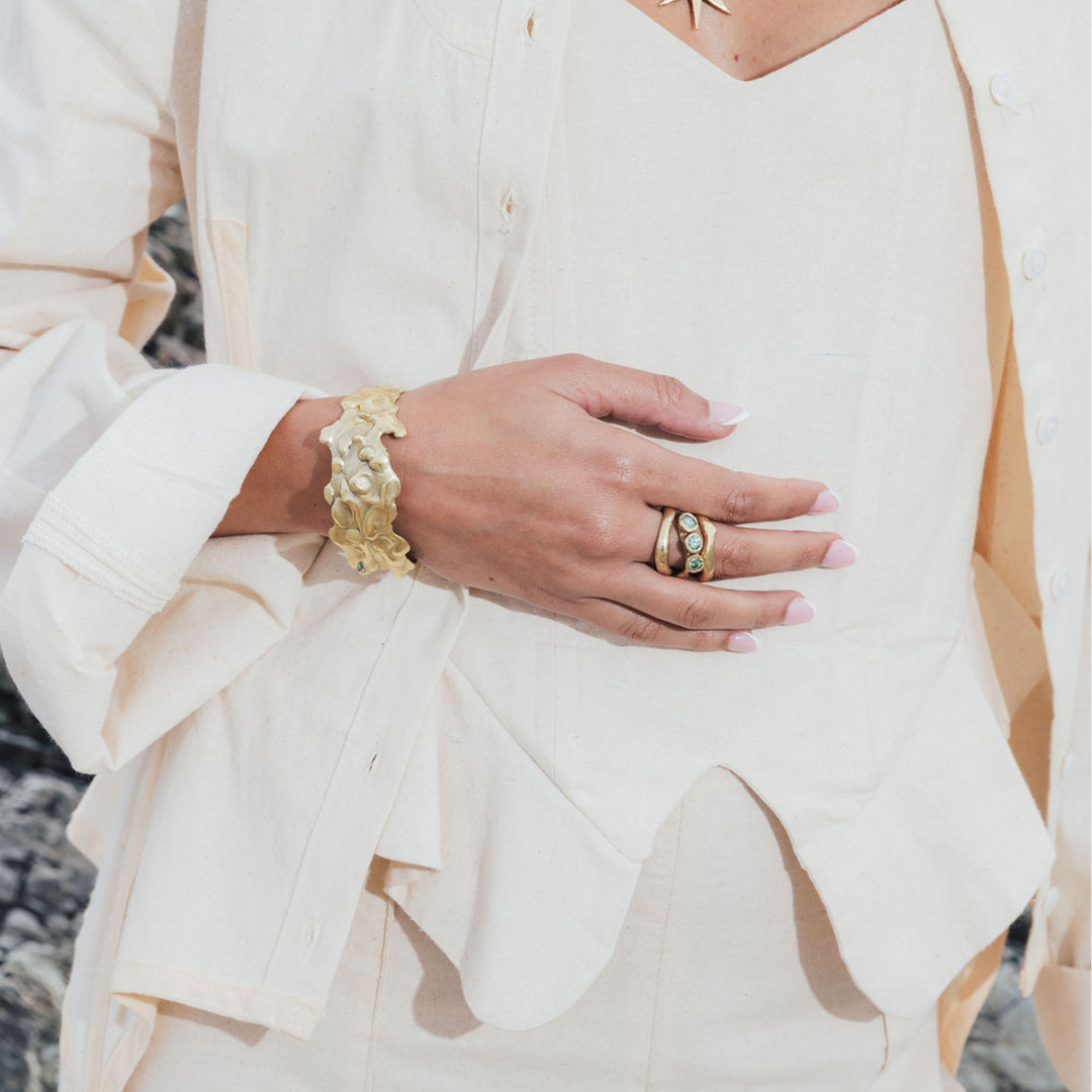 A close-up of a person's hand wearing a chunky gold textured cuff bracelet and multiple gold rings, styled with a cream-colored linen button-up shirt.