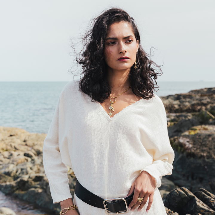 A woman with dark curly hair stands on a rocky shoreline wearing a loose white textured blouse with a V-neck, a black belt, and gold jewelry.