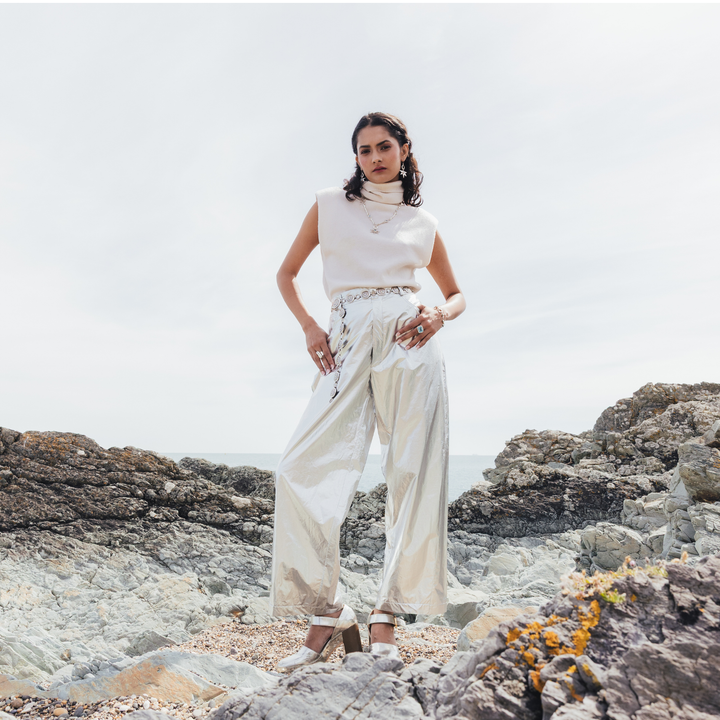 A woman stands on rocky coastal ground wearing a sleeveless white turtleneck top and shiny cream wide-leg pants with embroidered details, photographed outdoors against a pale sky and sea.