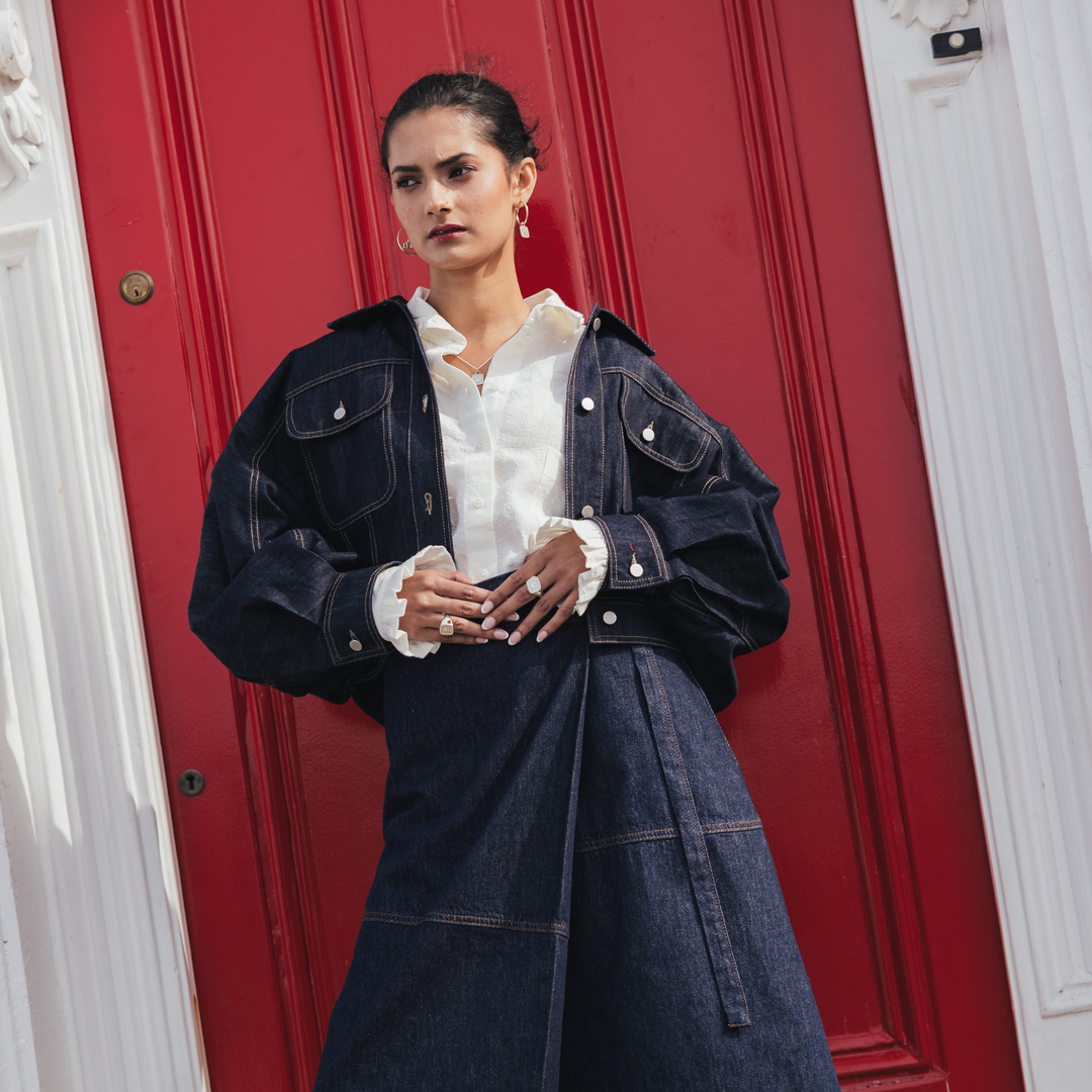 A model stands in front of a red door wearing an oversized dark indigo denim jacket over a white collared shirt, paired with a matching dark denim skirt.