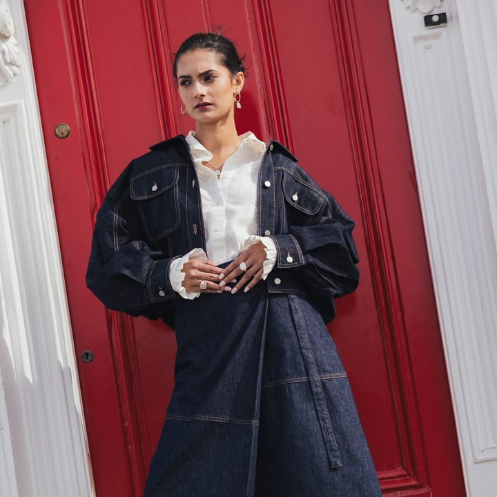 A model stands in front of a red door wearing an oversized dark indigo denim jacket over a white collared shirt, paired with a matching dark denim skirt.