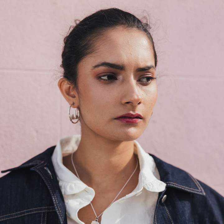 A close-up portrait of a woman wearing gold hoop earrings, a silver necklace, a white collared shirt, and a dark denim jacket against a soft pink wall.