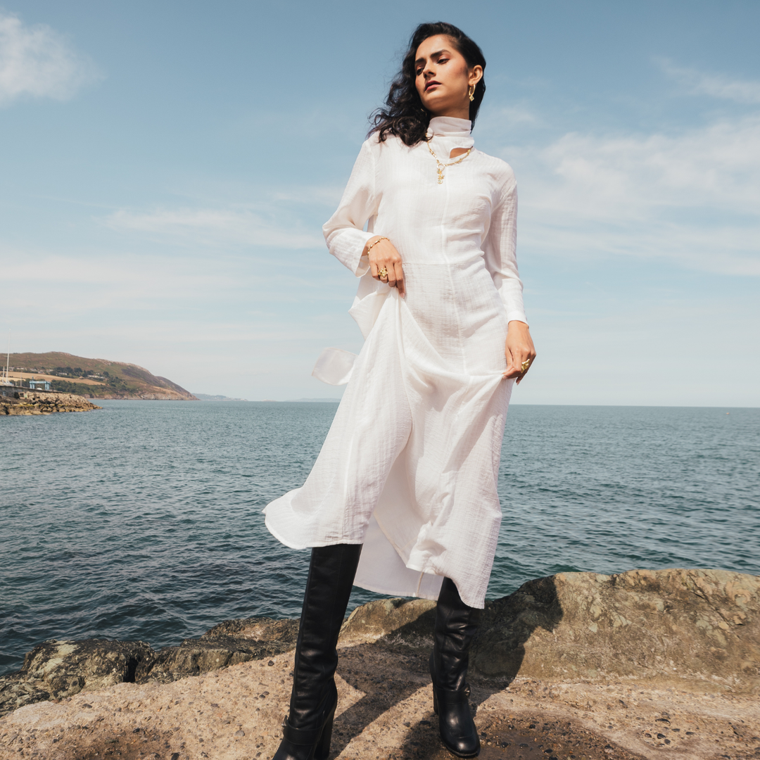 A woman stands on a rocky shoreline wearing a flowing white long-sleeve tunic dress with black knee-high boots, with the sea and sky behind her.
