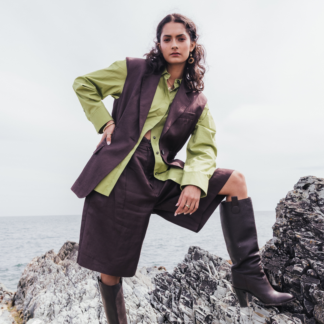 A model stands on rocky coastal ground wearing a sage green shirt, a dark brown structured vest, a matching dark brown skirt, and dark brown knee-high boots.