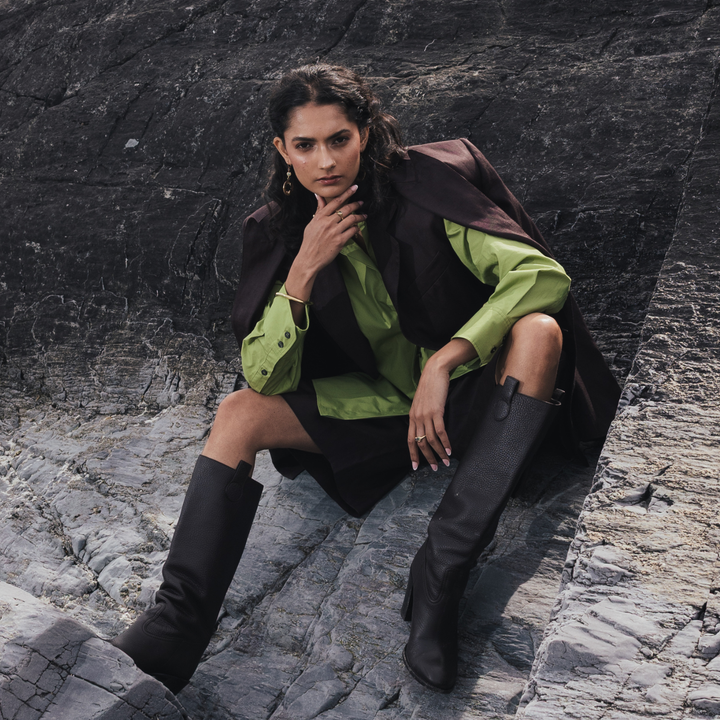 A model sits on a rocky outdoor surface wearing a dark brown sleeveless vest over a bright green shirt, paired with a black mini skirt and tall black boots.