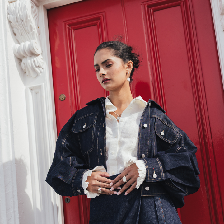 A woman wearing a dark indigo denim jacket over a white ruffled blouse, standing in front of a bright red door with white architectural molding on the left.