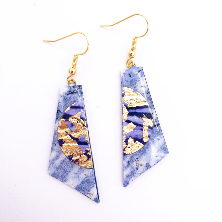 A pair of Cora Batik Textile Earrings in Lilac/Gold by Rothlú, featuring asymmetrical lilac stone-like pendants with gold leaf accents. These irregularly trapezoidal pendants are attached to hypo-allergenic gold plated hooks and combine elements of marbling and abstract artistry for a unique texture.