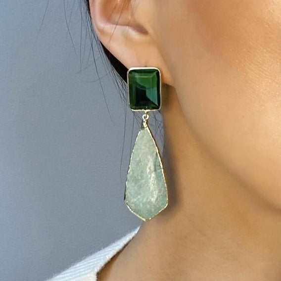 Close-up of a person wearing Aria V Cote d'Azur Earrings, showcasing a statement design with Amazonite stones. A large, square dark green gemstone sits at the top, while a teardrop-shaped light green stone dangles beneath. Set in 16 k gold brass, the earrings contrast beautifully with the person's skin against a neutral background.