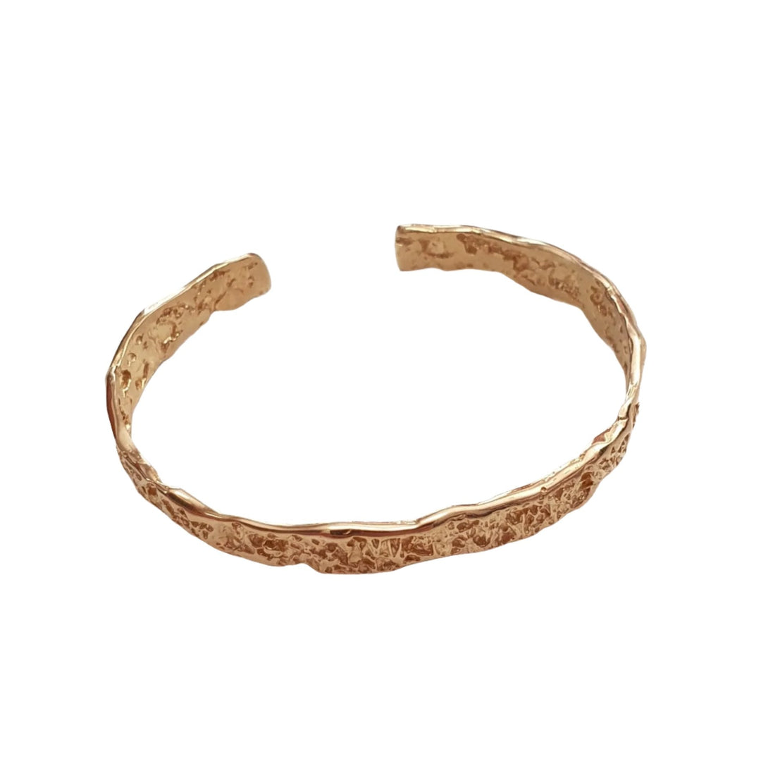 A textured, open-ended gold-plated bracelet is laid flat on a white surface. The Crackle Texture Bangle from Georgia Charal has a rugged, organic design with an uneven surface, giving it a handmade appearance.