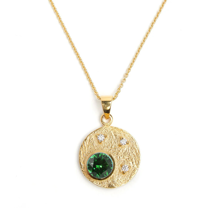 The Celeste Necklace in Gold & various gemstones by Gallardo & Blaine Designs features a round pendant adorned with a large green gemstone and three cubic zirconia stones, its textured surface and delicate chain adding elegance to any jewelry collection.