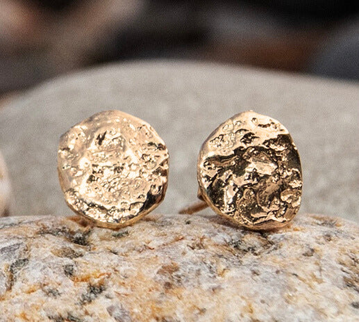 A close-up of textured, round 9ct Gold Dublin Bay Silver Studs by Ava Jane Jewellery, resting on a smooth, light-colored stone surface.