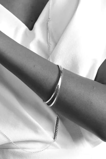 A black and white portrait of a person with a serene expression. They're dressed in a white, short-sleeved top, adorned with a long hand-forged necklace and a Lynsey de Burca Doon Bangle. Their hair is pulled back, and they are clasping their hands gently in front of them against a plain backdrop.