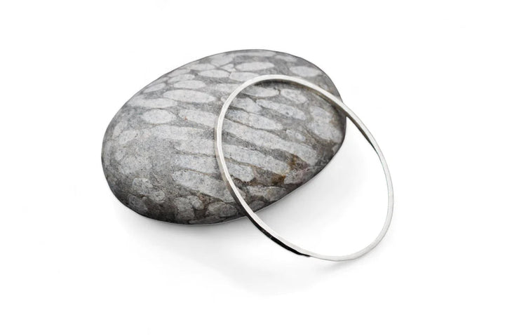 A hand forged, sterling silver Lynsey de Burca Doon Bangle rests on a smooth, oval-shaped gray stone with white lines and patterns against a plain white background.