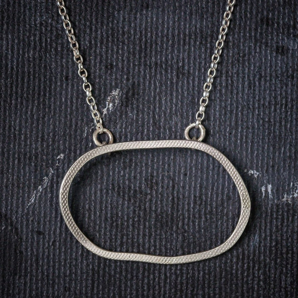 A textured, oval-shaped sterling silver pendant with a delicate chain. The open oval design features a subtle, hand-forged hammered finish. The Lynsey de Burca Drift Pendant Silver is displayed against a dark, textured background, evoking the rugged beauty of the Wild Atlantic Way.