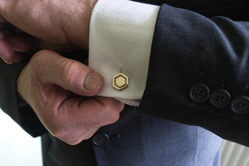A person is adjusting the cuff of their white dress shirt, which is adorned with Millett Wade Hexagon Cufflinks. They are wearing a dark suit jacket with three black buttons. The image focuses on the hands and the shirt cuff.