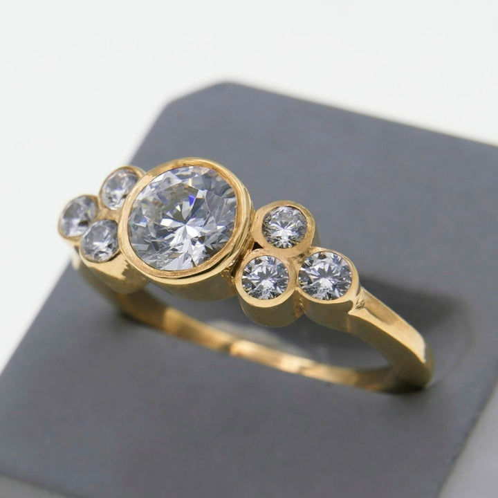 A stunning gold Sionna - Seven Stone Engagement Ring by Miriam Wade featuring a central 1 carat lab grown diamond flanked by three smaller round diamonds on each side. The ring is displayed on a gray stand against a white background.