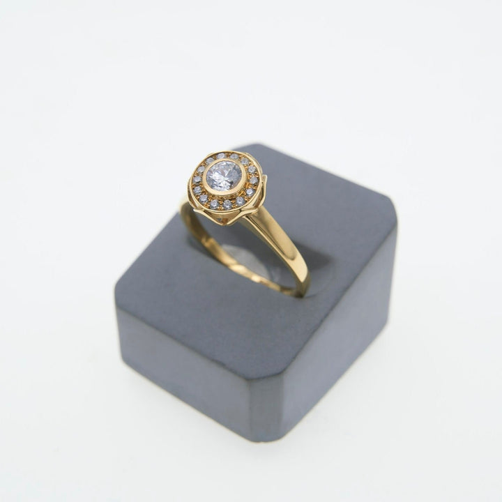 A gold Éirne - Diamond Halo Engagement Ring by Miriam Wade with a round, central clear gemstone set in a halo of smaller lab-grown diamonds. The ring is displayed on a dark gray square pedestal against a white background.