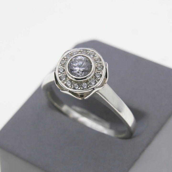 A silver ring featuring a central round diamond surrounded by two concentric circles of smaller, lab-grown diamonds. The Éirne - Diamond Halo Engagement Ring by Miriam Wade, reminiscent of a delicate floral pattern, is displayed on a dark gray cushion against a light background.