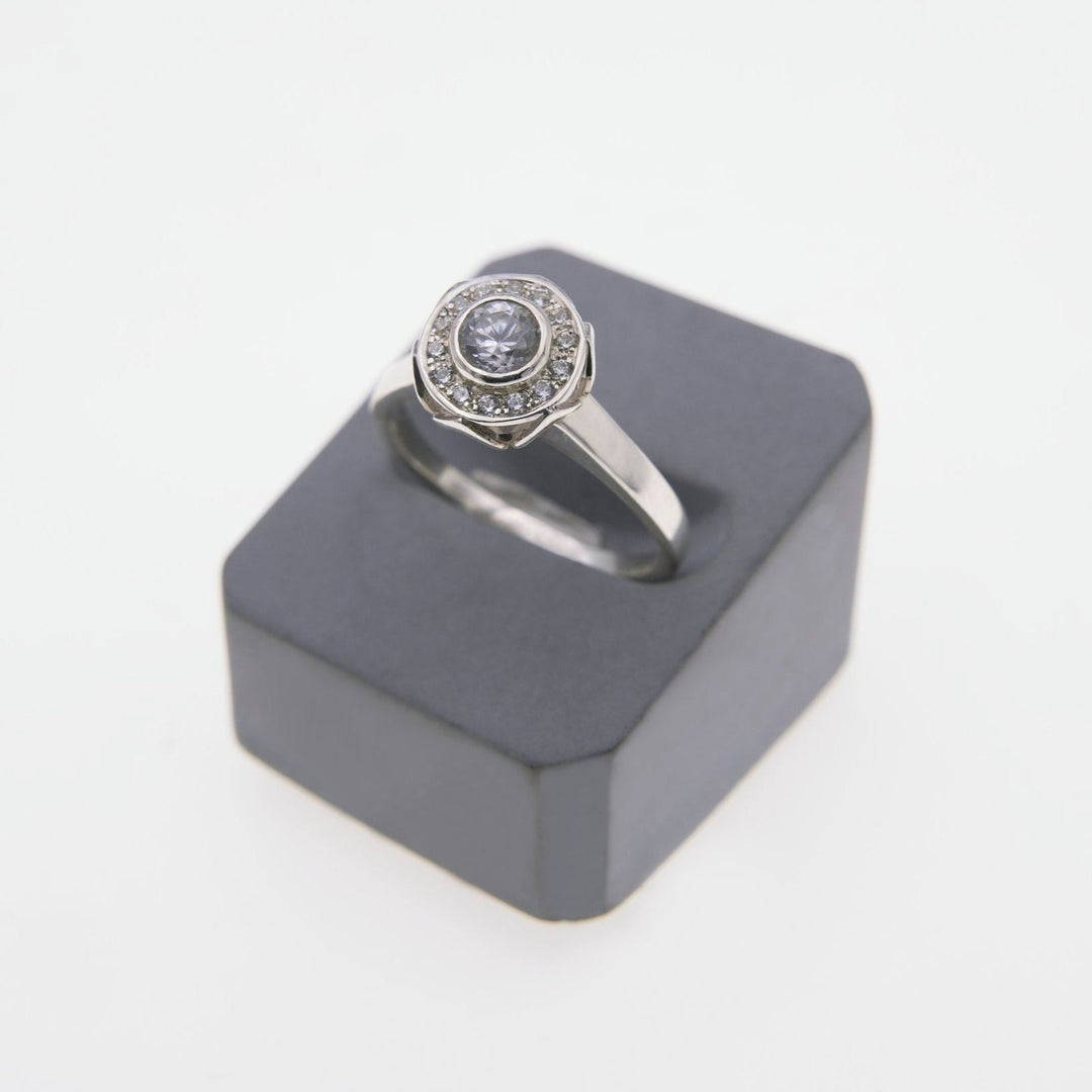 A **Miriam Wade Éirne - Diamond Halo Engagement Ring** with a round, diamond-encrusted setting is displayed on a dark grey, hexagonal stand. The ring's central stone is surrounded by smaller stones in a gold diamond halo engagement ring style, creating a sparkling and elegant appearance. The background is plain white.