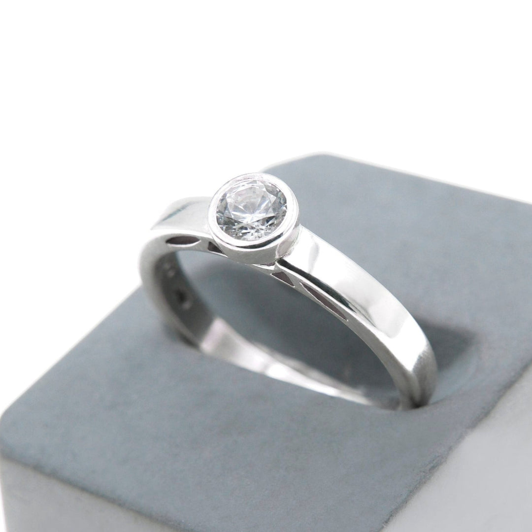 A close-up view of a gold solitaire ring with a single round lab grown diamond set in a bezel setting, placed on a blue-gray, square-angled stand against a white background. The Inagh - Solitaire Engagement Ring's polished surface and minimalist design by Miriam Wade are showcased.