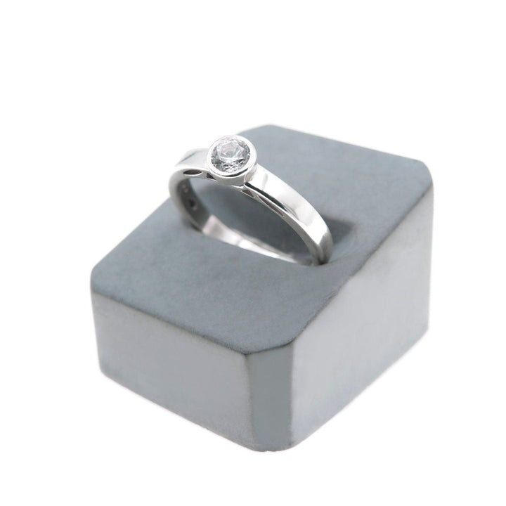 A silver **Inagh - Solitaire Engagement Ring** by **Miriam Wade** with a single round lab grown diamond set in a raised bezel setting is displayed atop a gray velvet hexagonal ring box. The background is plain white, drawing attention to the ring.