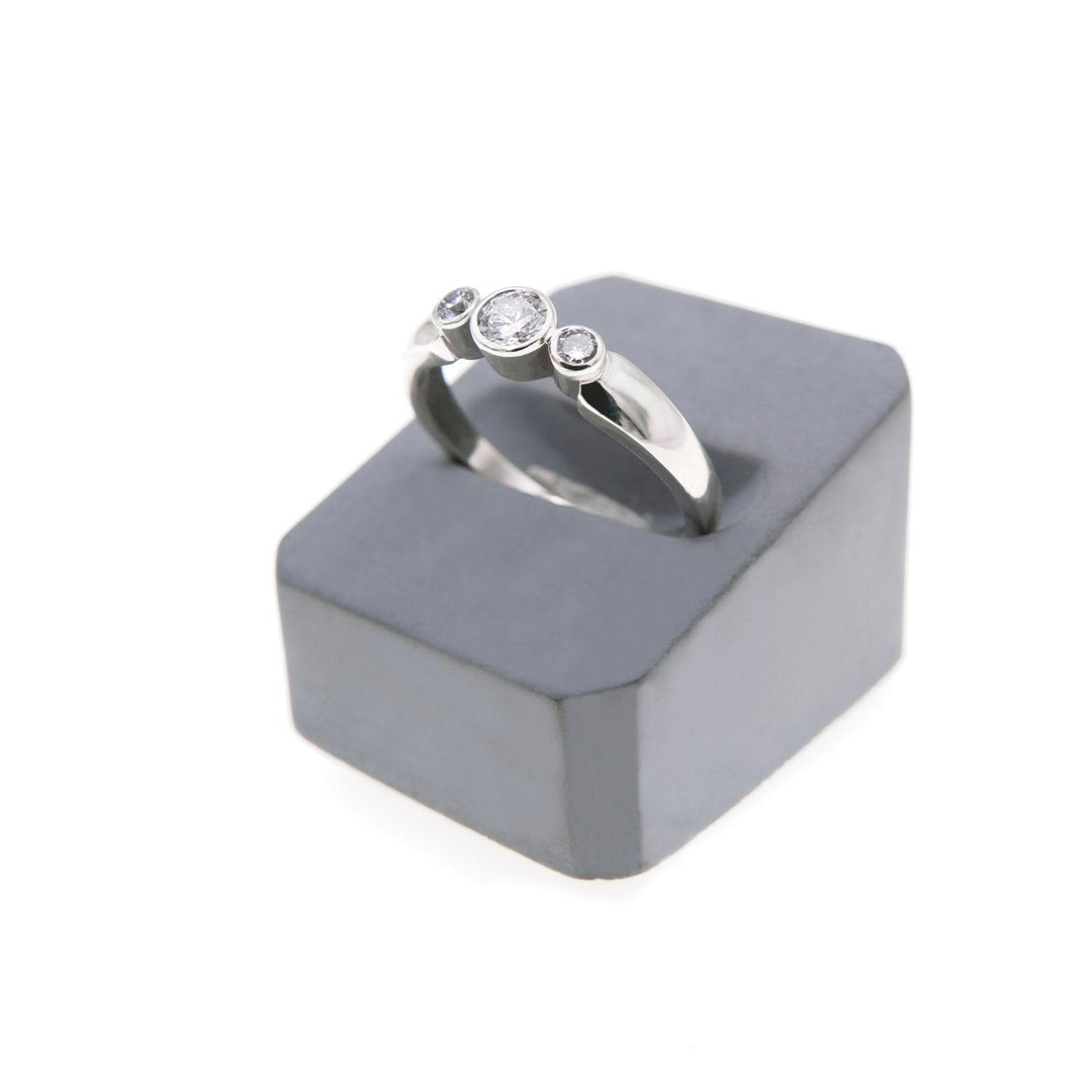 A handcrafted silver ring featuring lab grown diamonds is displayed on a gray, rectangular stand. The Síleann - Three Stone Engagement Ring by Miriam Wade has the largest diamond in the center, flanked by two smaller diamonds on either side.