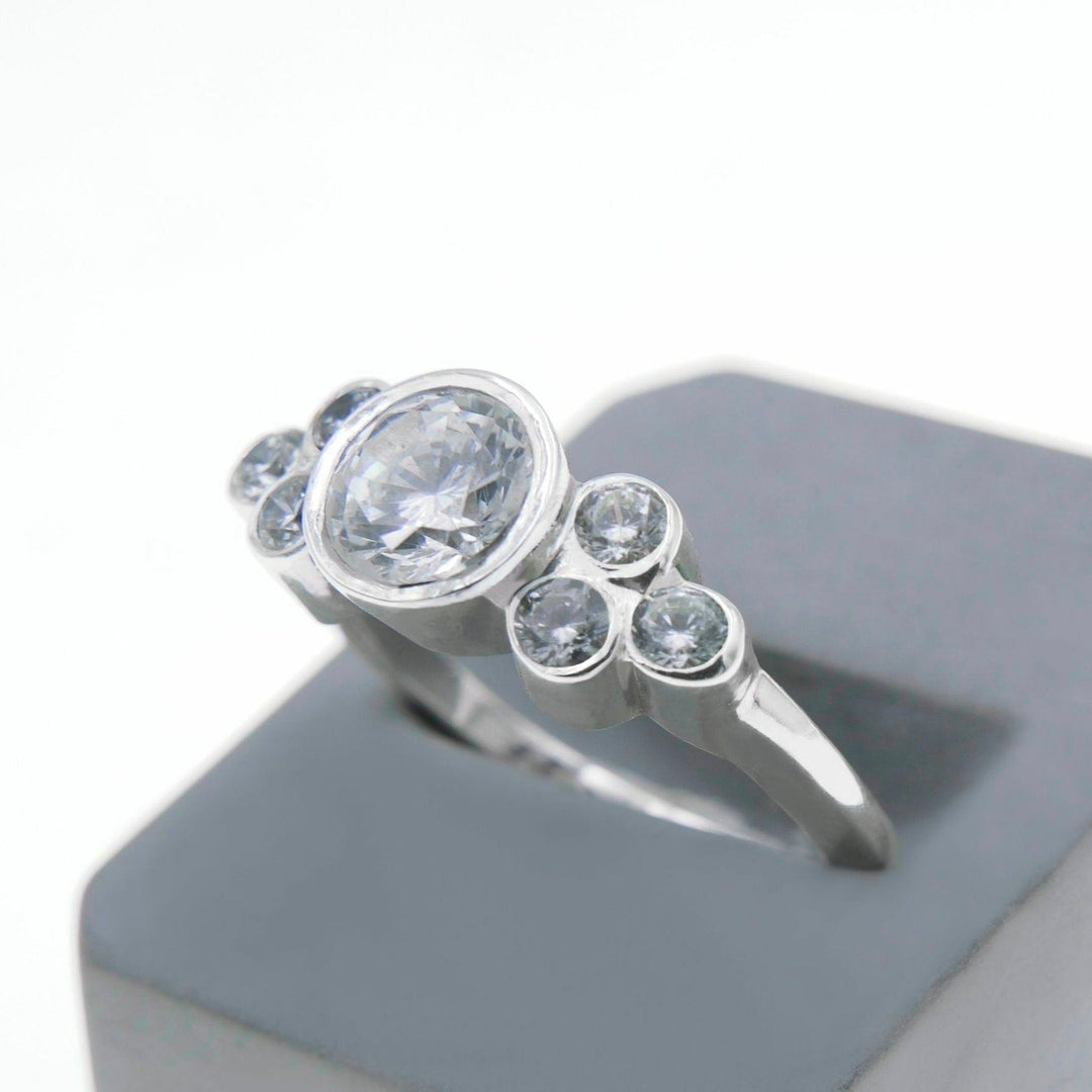 A Miriam Wade Sionna - Seven Stone Engagement Ring with a 1 carat diamond at its center and four smaller lab grown diamonds set in descending order on each side is displayed on a gray, angular pedestal against a plain white background.