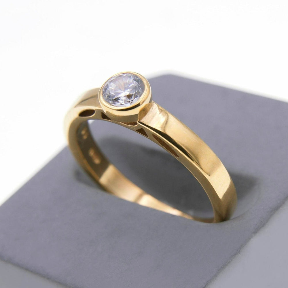 The Inagh - Solitaire Engagement Ring by Miriam Wade with a single round lab-grown diamond set in a bezel setting is displayed on a white and gray platform. The ring's design features a polished, smooth band, elegantly curving around the finger. The diamond captures light, adding sparkle to the overall look.