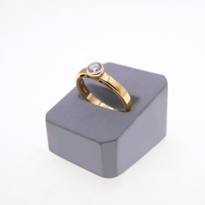 A gold solitaire ring with a round, clear gemstone set in the center is displayed on a gray, rectangular stand against a white background. The Inagh - Solitaire Engagement Ring by Miriam Wade has a sleek, modern design, and the lab grown diamond is prominently showcased.