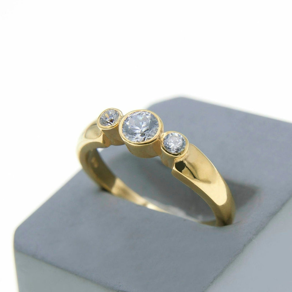 A Miriam Wade Síleann - Three Stone Engagement Ring adorned with lab-grown diamonds of varying sizes is placed on a light grey, padded display stand. The central diamond, the largest, is flanked by smaller ones on each side. The ring, crafted from handcrafted precious metals, is shown at a slight angle to highlight its design.