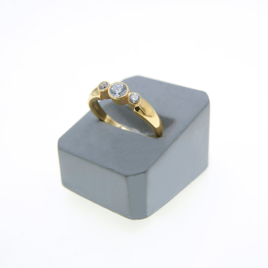 A Síleann - Three Stone Engagement Ring by Miriam Wade is displayed on a gray, rectangular stand. The ring features a large central diamond, flanked by two smaller lab-grown diamonds on either side. The background is white, making the handcrafted precious metals and gems stand out beautifully.