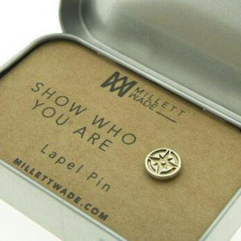 A small silver lapel pin with a star design is displayed inside an open tin container. The cardboard insert in the container reads "SHOW WHO YOU ARE, Compass Lapel Pin, Millett Wade." Evoking a sense of adventure and travel, the container with its hinged lid is partially visible.