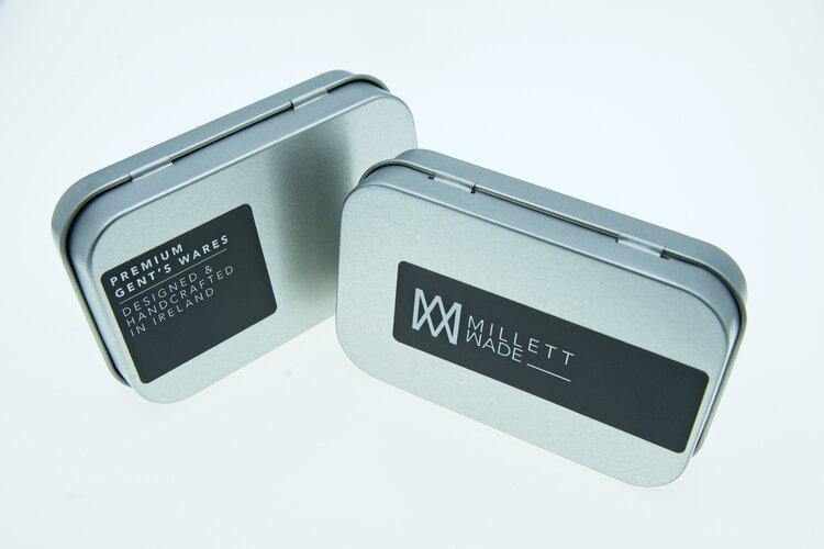 Two rectangular metallic tins with dark labels. The left tin label reads "Premium Gentlemen's Wares Designed and Developed in Ireland." The right tin label features the "Millett Wade" brand logo and name. Both tins have smooth, rounded edges and a sleek design, ideal for holding a Millett Wade Compass Lapel Pin.