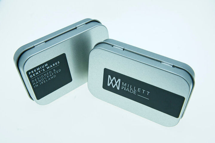 Two rectangular metal tins are displayed against a white background. Both tins have black labels; one reads "PREMIUM GENTS WARES, DESIGNED AND CREATED IN IRELAND," and the other reads "Millett Wade" with a logo. Inside, you'll find hand-finished Hexagon Cufflinks crafted from solid brass.