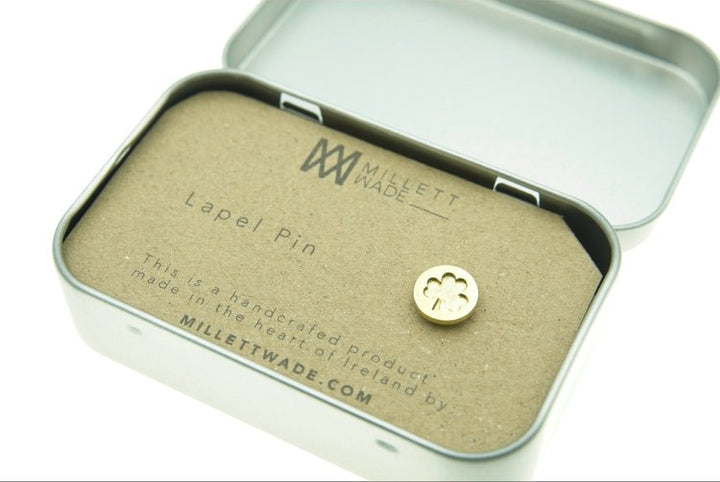 A small metallic lapel pin shaped like a four-leaf clover is displayed inside an open metal tin container. The card inside the tin reads "Millett Wade Shamrock Lapel Pin" and details that it is handcrafted in Ireland, boasting high quality metal lacquer, also providing the website "MILLETTWADE.COM.