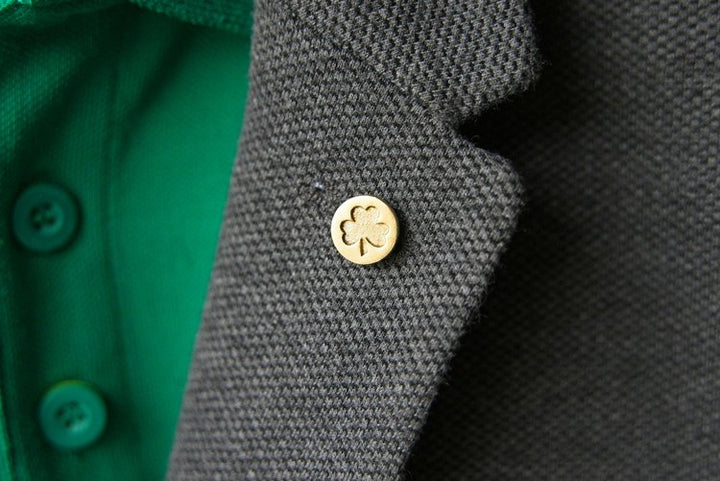 Close-up of a gray textured blazer featuring a gold button adorned with a four-leaf clover design. A solid brass lapel pin, the Shamrock Lapel Pin by Millett Wade, adds an extra touch of sophistication, while the blazer is worn over a green shirt with buttons, creating a striking contrast.