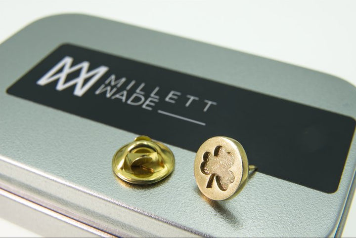 A Shamrock Lapel Pin is displayed on a metallic case with the logo and text "Millett Wade." The lapel pin features a four-leaf clover design. Hand-finished to perfection, the case is silver with a black label.