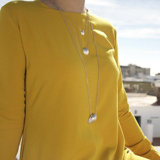 A person wearing a bright yellow long-sleeved top is shown from the shoulders to the waist. They are adorned with three silver chains of varying lengths, one of which is a gold-plated Jennifer Kinnear Cockle Shell Necklace Large. The background features a blurred urban setting under a clear sky.