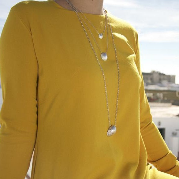 A person in a long-sleeved, yellow top is wearing two silver chains of different lengths. One sterling silver pendant is a small disc, and the other is a larger, slightly teardrop-shaped piece from the Cockle Shell Necklace Medium - Silver by Jennifer Kinnear. The background shows a sunny, urban outdoor scene with buildings and a blue sky.