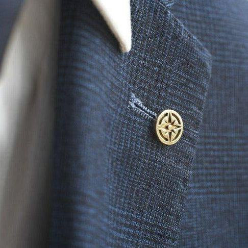 Close-up of a navy blue textured suit jacket with a Millett Wade Compass Lapel Pin, symbolizing adventure and travel. The background is blurred, highlighting the jacket's fabric and the custom pin detail. The edge of a white shirt is visible under the jacket.