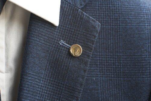 Close-up of a person wearing a dark blue checked suit jacket with a white shirt. The jacket lapel is adorned with a Millett Wade Harp Lapel Pin featuring a small, round, gold harp emblem. The pin, resembling fine hand-finished brass items, is fastened near the buttonhole.