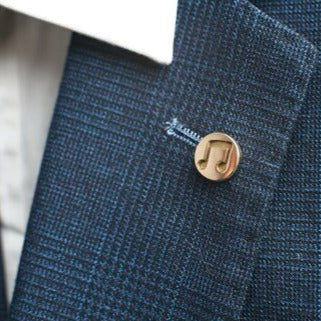 Close-up of a custom brass Millett Wade Music Note Lapel Pin attached to a blue textured blazer. The gold-colored pin stands out against the fabric, and the background shows the blurred shirt collar and part of the shirt in light colors.