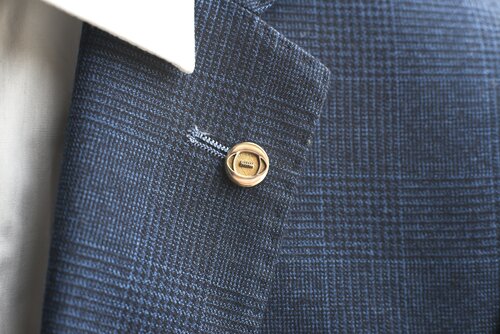 Close-up image of a blue business suit jacket with a subtle checkered pattern. The focus is on a beige, round button sewn onto the jacket, beside which sits an elegant brass Rugby Lapel Pin by Millett Wade. The edge of a white dress shirt collar is also visible in the background.