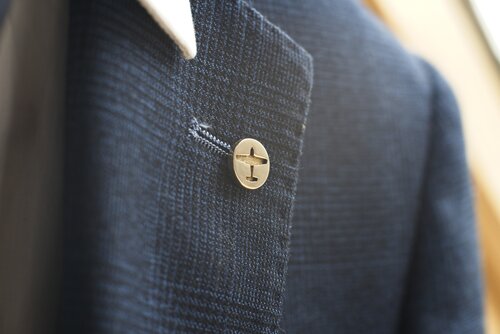 Close-up of a dark blue blazer with a white dress shirt underneath. The blazer features a buttonhole on the lapel with a custom brass pin inserted, showcasing the Millett Wade Plane Lapel Pin, displaying a small circular metal piece with a cross design engraved on it. The fabric has a textured weave.