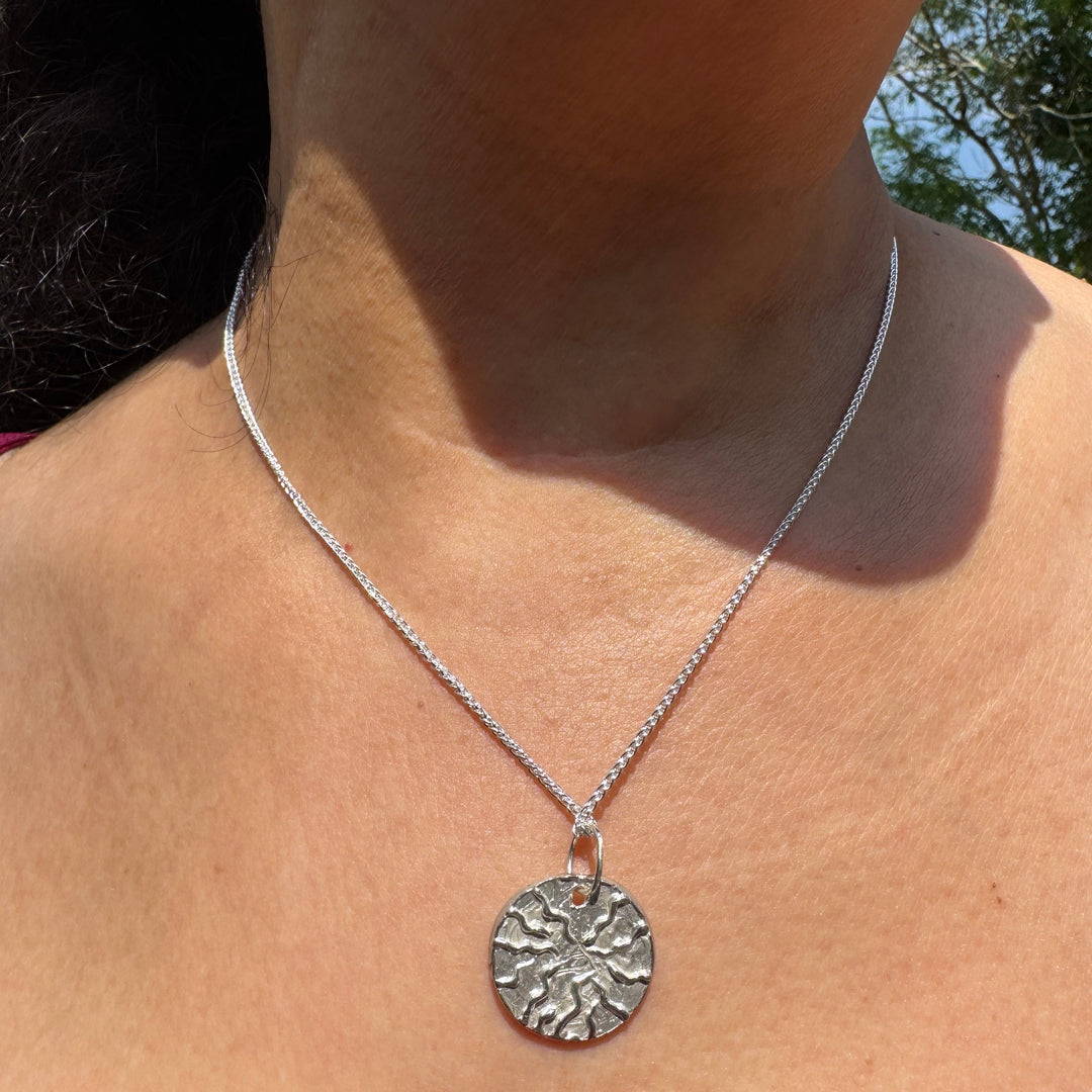 A person wears the Dara Pendant by Soufana Attia—a silver, textured round pendant symbolizing strength—against bare skin and a magenta off-shoulder top, with green foliage in the background.