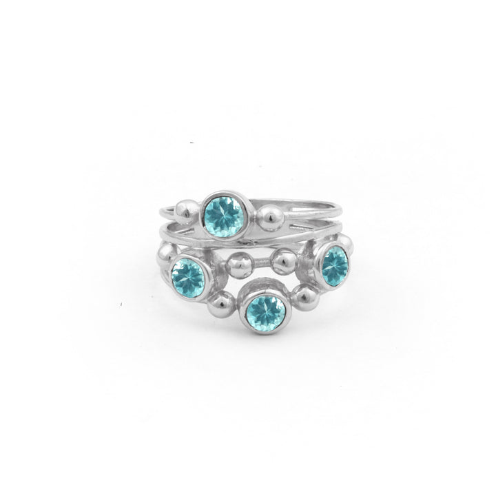 The Justine Ring in Apatite by Watermelon Tropical is a sterling silver ring featuring a unique, layered wrap design adorned with six evenly spaced, round apatite gemstones. The gemstones are set in circular bezels interspersed with small silver beads. The ring has a modern and elegant look against a white background.