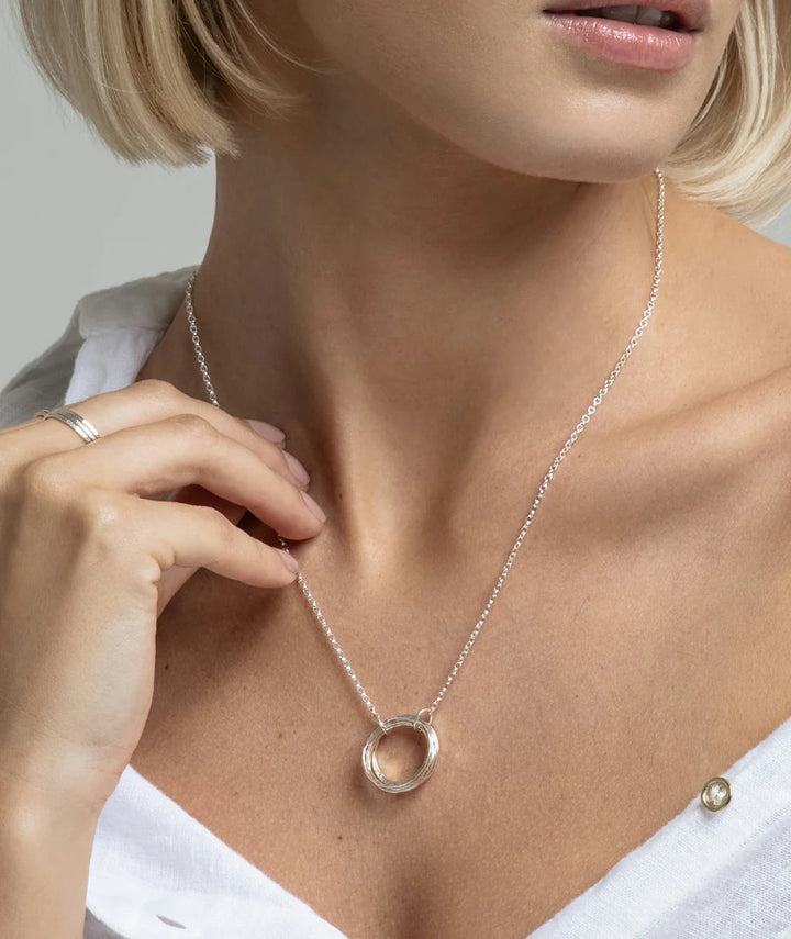 A close-up image of a person wearing Lynsey de Burca's Doorus Mix Pendant, a sterling silver necklace with a circular, hand-forged pendant. They have a short blonde bob haircut and are dressed in a white top. The person's hand is touching the Doorus Mix Pendant, and they are also wearing a silver ring on their index finger.