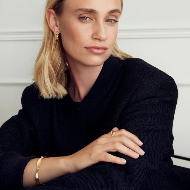 A woman with shoulder-length blonde hair in a black top poses with arms crossed, showcasing Watermelon tropical's Dora Bangle along with hoop earrings and a ring, against a white background.