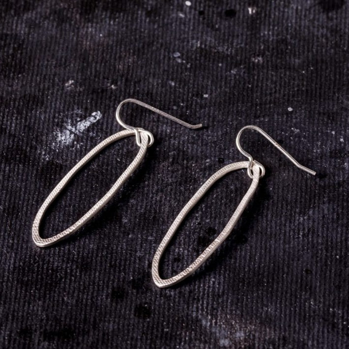 A pair of hand-forged, sterling silver oval-shaped Drift Drop Earrings with a minimalist design by Lynsey de Burca, displayed on a dark fishing net rope texture background.